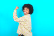 © Roquillo - Portrait of powerful cheerful young girl with afro hairstyle wearing floral shirt over blue background showing muscles.