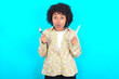 © Roquillo - hungry young girl with afro hairstyle wearing floral shirt over blue background holding in hand fork knife want tasty yummy pizza pie