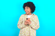 © Roquillo - young girl with afro hairstyle wearing floral shirt over blue background holding a smartphone and looking sideways at blank copyspace.