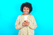 © Roquillo - young girl with afro hairstyle wearing floral shirt over blue background enjoys distant communication, uses mobile phone, surfs fast unlimited internet, has pleasant smile, makes shopping online,