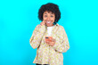 © Roquillo - Joyous young girl with afro hairstyle wearing floral shirt over blue background poses with mobile phone device, types text message on modern smartphone, watches funny video during free time,