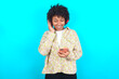 © Roquillo - Happy young girl with afro hairstyle wearing floral shirt over blue background feels good while focused in screen of smartphone. People, technology, lifestyle