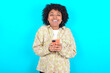 © Roquillo - Excited young girl with afro hairstyle wearing floral shirt over blue background holding smartphone and looking amazed to the camera after receiving good news.