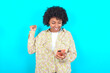 © Roquillo - young girl with afro hairstyle wearing floral shirt over blue background holding in hands cell and rising his fist up being excited after reading good news.