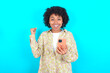 © Roquillo - Portrait of young girl with afro hairstyle wearing floral shirt over blue background holding in hands cell celebrating