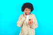 © Roquillo - young girl with afro hairstyle wearing floral shirt over blue background holding in hands cell reading sms using new app 5g