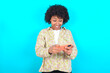 © Roquillo - Portrait of an excited young girl with afro hairstyle wearing floral shirt over blue background playing games on mobile phone.