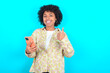 © Roquillo - Portrait of young girl with afro hairstyle wearing floral shirt over blue background using and texting with smartphone  happy with big smile doing ok sign, thumb up with fingers, excellent sign