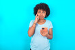 © Roquillo - Young woman with afro hairstyle wearing blue T-shirt over blue background holding gadget while sticking out tongue