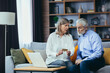 © Liubomir - Senior couple, gray-haired retirees sitting on sofa together at home, wife explains to husband how to use credit card for online shopping in online store