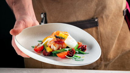 The chef is holding a plate of salad in his hands. Warm salad with shrimp, tomatoes, avocado and arugula, original cuisine, close up