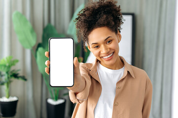 African American beautiful positive girl with curly hair in casual wear, standing at home, holding a smartphone in her hand with blank white screen for advertising, looking at camera, smiles happily