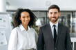© Kateryna - Portrait of two successful multiracial colleagues, company co-founders, directors, managers, a caucasian man and an African American woman, standing in a modern office, looking at camera smiling