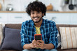 © kucherav - Young Asian man using mobile phone, chatting online sitting at home on the couch. Portrait of Indian male holding smartphone looks at camera and smiles friendly