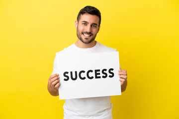 Wall Mural - Young handsome caucasian man isolated on yellow background holding a placard with text SUCCESS with happy expression