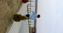 Curly-haired Girl With Red Hair Runs Along The Edge Of The Lake Along The Thuja And Turns Onto The Bridge. Rear View, Wiring Movement With Steadicam. Vertical View