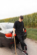 © alones - Fashionable stylish young hipster man in black clothes with a longboard stands near the car on the road near the cornfield. Active weekend and travel