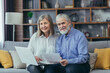 © Liubomir - Senior gray-haired married couple man and woman sitting together on sofa at home, looking at camera and smiling, paper work with laptop