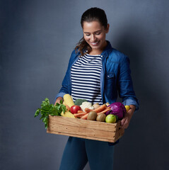 Sticker - Everyone says she is so down to earth. Studio shot of a beautiful young woman holding a wooden crate full of fruit and vegetables against a blue background.