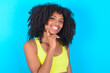 Carefree successful young woman with afro hairstyle in sportswear against blue background touching jawline gazing camera tilting head grinning white teeth delighted. Dental care concept.