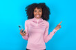 © Jihan - Astonished young woman with afro hairstyle in technical sports shirt against blue background  holding her telephone and pointing with finger aside at empty copy space