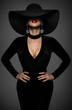 Sexy Fashion Woman in Big Hat and Black Dress. Beauty Model with Curve Body Shapes in Seductive Bodycon Gown. Mysterious Elegant Lady over Dark Gray Background. Women hidden Face and Red Lips