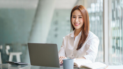 Wall Mural - Beautiful young Asian businesswoman sitting working with a laptop at the office. Looking at camera.