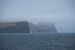 Passenger ferry Norröna Norroena passing sharp and steep rocks and cliffs of Faroese coast of Faroe Islands with mountains, waterfalls and tide in magic mystical foggy day with low hanging clouds