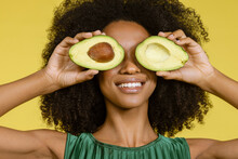 Smiling Young Woman Covering Eyes With Avocados Against Yellow Background