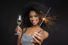 Smiling Young Woman Holding Champagne Flute And Sparkler Against Black Background