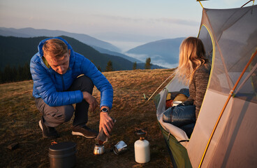 Wall Mural - Morning in summer tent camp in the fresh air. Man preparing breakfast for his girlfriend. Morning fog over mountain hills on the background. Concept of hiking, camping and relationships.