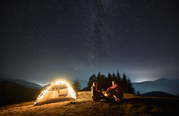 Wall Mural - Woman with guitar in her hands sitting opposite man by campfire near their light shining tent. Romantic evening with loved one outdoors under starry sky in the mountains. Concept of night camping.