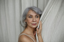 Thoughtful Senior Woman With Gray Hair Touching Curtain At Home