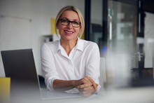 Smiling Businesswoman Wearing Eyeglasses Sitting With Laptop At Desk In Office