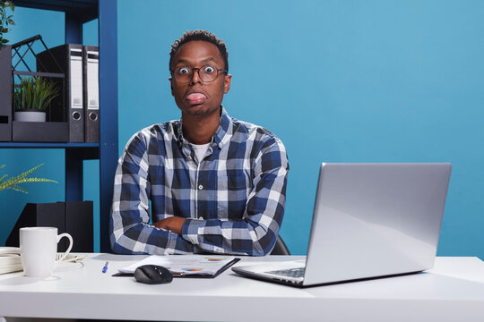 Funny and goofy office working person making amusing playful face while sticking out tongue. Childish and dumb young adult man with dullard face expression while in company workspace.