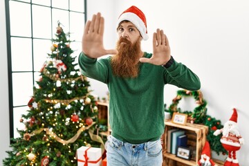 Sticker - Redhead man with long beard wearing christmas hat by christmas tree doing frame using hands palms and fingers, camera perspective