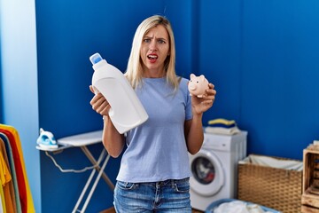 Poster - Beautiful woman doing laundry holding detergent bottle and piggy bank clueless and confused expression. doubt concept.