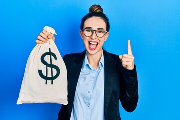 Poster - Young hispanic girl wearing business clothes holding dollars bag smiling with an idea or question pointing finger with happy face, number one