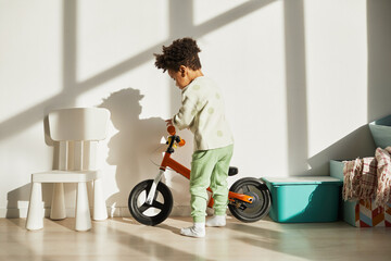 Wall Mural - Minimal full length portrait of African American little boy riding balance bike at home in sunlight, copy space