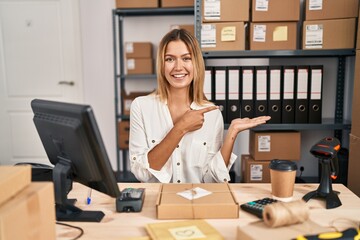 Canvas Print - Young blonde woman working at small business ecommerce amazed and smiling to the camera while presenting with hand and pointing with finger.