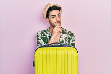 Wall Mural - Young hispanic man wearing summer style and holding cabin bag smiling looking confident at the camera with crossed arms and hand on chin. thinking positive.