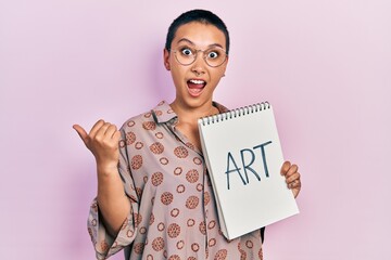 Poster - Beautiful hispanic woman with short hair holding art notebook pointing thumb up to the side smiling happy with open mouth