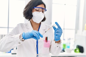 Canvas Print - Young latin woman wearing scientist uniform and medical mask holding vaccine at laboratory