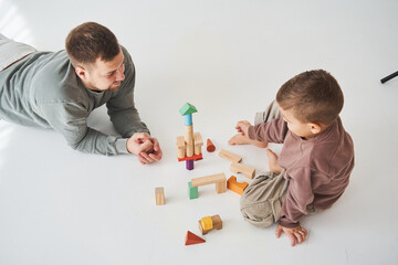 Caring dad helps his son to play on the floor on white background. Father and child build tower of colorful wooden bricks and have fun together.