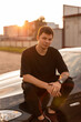 © alones - Fashionable young man model in stylish black summer clothes sits on the car at sunset