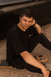 © alones - Hipster handsome man model in black fashionable clothes sits and rest on a longboard