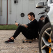 © alones - Hipster handsome man in black t-shirt, jeans and sneakers uses a smartphone and sits on longboard near a car. Summer weekend