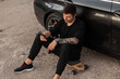 © alones - Fashionable handsome hipster man in black clothes with a smartphone sitting and rest on a longboard near his car. Outdoor activities and weekend