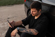 © alones - Happy young handsome hipster man in a black T-shirt with a smartphone sits near a car and uses the phone