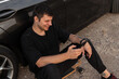 © alones - Fashionable happy hipster man in black clothes using a smartphone and smiling, sitting on a longboard near car